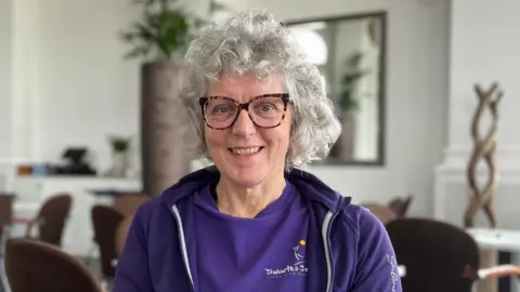 Juliette has short, curly grey hair and is seated indoors in a bright, modern café-style room. She is wearing a purple zip-up jacket over a matching purple T‑shirt. Behind her are brown chairs, white tables, tall white walls, and several potted plants, including one large plant positioned directly behind their seat.