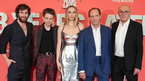 Four actors and Danny Boyle pose in a row, standing against a promotional red board featuring the title of the film 28 Years Later.