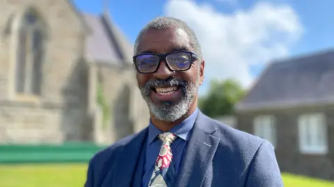 George Hargreaves, who has a grey beard, smiles. He wears dark-rimmed glasses and a blue suit and colourful tie. 