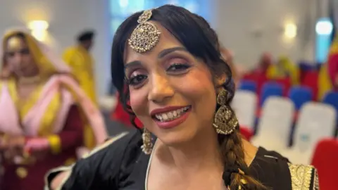 A woman with dark hair, a dark top and large gold earrings and a gold head decoration