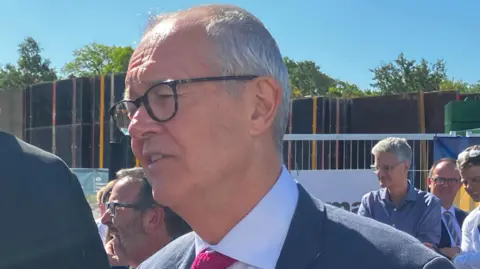 Lord Patrick Vallance is side on to the camera, has grey hair, dark framed glasses, is wearing a navy suit and red tie.