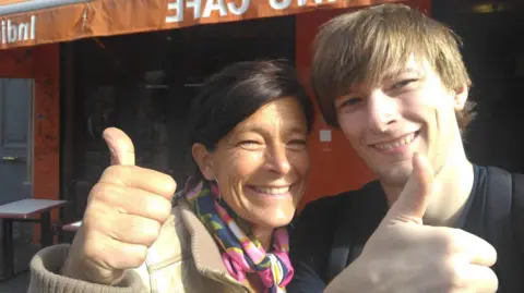 A man and woman with brown hair smile and give a thumbs up to the camera outside a cafe with orange bunting