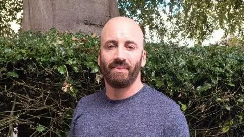 A man in a grey T-Shirt is standing in front of a green hedge. He is bald and has a brown beard. He is smiling at the camera.