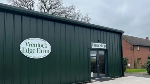 Ellen Knight/BBC Photograph of the farm shop since its reopening. It is a large, single-storey building, clad with dark green corrugated metal. A large, oval, white sign is on the wall, which reads 'Wenlock Edge Farm' in green writing, with 'Artisan products, bacon sausages and ham' written around the edge. 