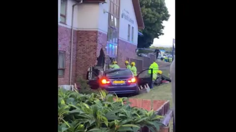 SGM A blue BMW car with its doors open and its rear headlights on, halfway through a brick building. Firemen in hi-vis jackets surround the car.