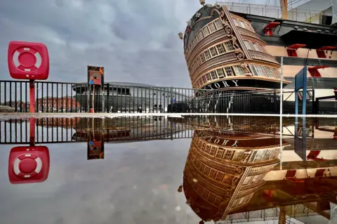 Walking Tractor An image of the stern of the HMS Victory in its dock with a perfect reflection in water. A life ring in its housing is on the left of the shot, equally reflected. Railings and a building sit in the background.