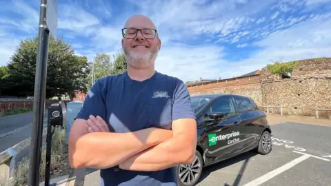 Stuart Woodward/BBC Andy Burlinson, standing next to an electric car in Priory Street car park in Colchester