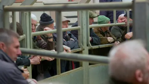 Men in coats and flat caps are leaning on a metal gate.