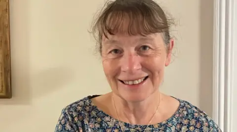Northumberland County Council 62 year old Barbara Hubbard with brown wispy hair stands smiling warmly in her home. She's wearing a patterned blue top and standing in front of the neutral walls of her home. 