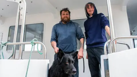 Captain Kimmy Reid with his crew member Aaron, and Bruce the dog. They are standing on a white boat.