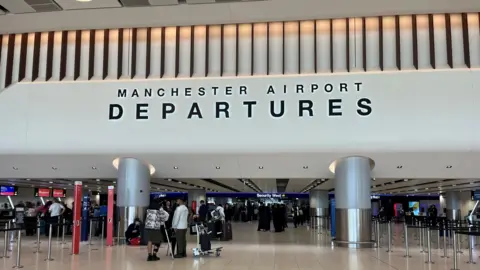 Inside Manchester Airport's departures check-in showing passengers milling about and the security section in the background.