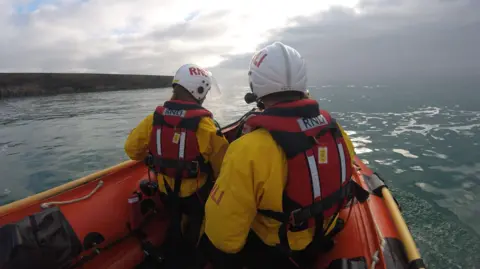 RNLI/Newhaven Two RNLI volunteers stand in Newhaven's D class lifeboat near breakwater during training. They are wearing red and yellow kit with the RNLI logo on it and standing looking out to sea.