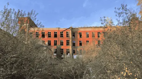 Irish Linen Centre & Lisburn Museum One of the site's buildings can be seen amongst overgrown trees. The orange brick of the building can be seen alongside numerous broken windows. This is amongst a blue sky. The image was taken in November 2024.