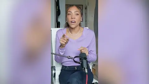 A woman sits in front of a microphone talking to someone across from her. She wears a purple top and dark blue jeans