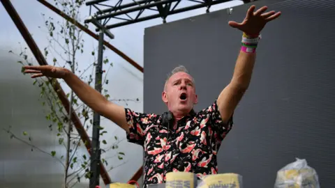 Getty Images DJ Fatboy Slim plays on stage at Glastonbury Festival. He is stood in front of a darkened screen and has his hands in the air with his mouth open. He is an older man with short grey hair and is wearing a black shirt with orange and white koi fish on it. He has a pair of headphones around his neck.