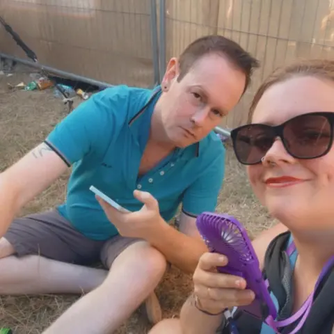 Supplied Man with short brown hair, blue top and grey shorts and Savannah Wright with brown hair glass and holding a fan. They are both sat on a floor in front of a fence.