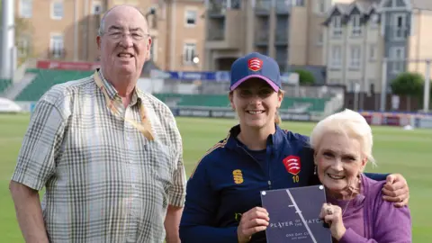 Dave and Pam Letch on the pitch at the County Ground. Dave is standing up while wearing glasses and a chequered shirt. Eva Gray is wearing a navy cricket strip and hat, and has her arm around Pam's shoulders. Pam is wearing a purple outfit and presenting Gray with a player of the match award.