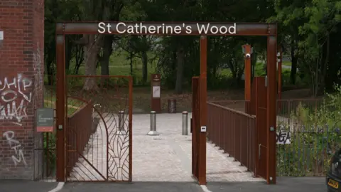 A red metal gated entranced to a park area. There are three silver bollards and established trees and grass area in the distance.