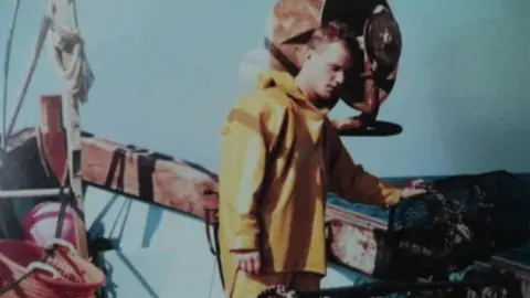 Steve Barrett A young Steve Barrett in yellow fishing overalls, stood on a fishing vessel.