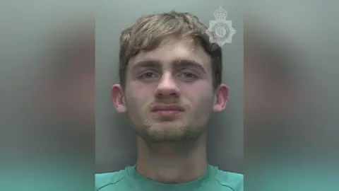 Humberside Police A close up picture of a Liam Best, a young man, in a police custody picture. Behind him is a solid grey background. He wears a teal t-shirt and has facial hair of a moustache and beard. He has light brown hair. 