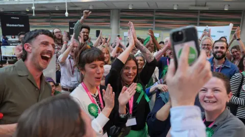 PA Media A group of men and women wearing Green Party badges laughing, smiling, clapping and posing for photos