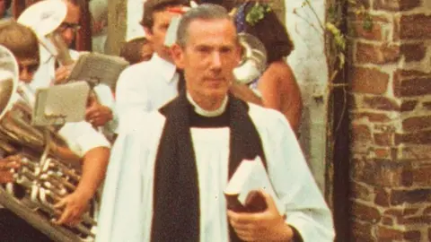 National Trust A photograph of a vicar, holding a prayer book is in a street in Mevagissy in front of a band 