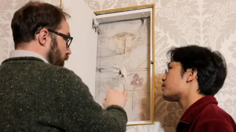 Durham University Andrew Ferrara and a student pointing at a cut-out in a wall which reveals the wall painting. Ferrara has black glasses and brown hair and a thick green jumper. The student is wearing a red shirt and is wearing glasses and has black hair. The wall painting is of a red flower with a green stem on a wall. There are red lines above and below and in the middle of the flower. 