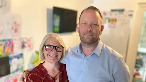 Duncan and Jo Sargent stood together. They are both looking at the camera. Duncan is wearing a light blue shirt and Jo is wearing a red patterned shirt.