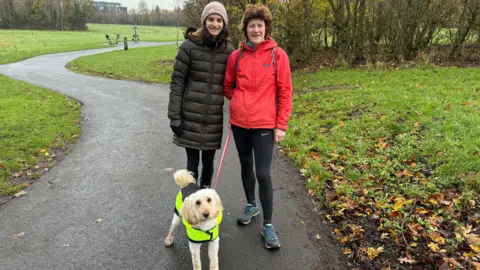 Two women stand in a park with their golden coloured dog. One woman is wearing a red rain coat and leggings, the other a dark coloured coat and beige hat. Around them is grass with fallenm leaves, and behind them a row of trees.