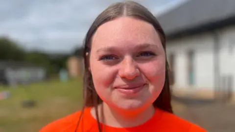 Rachel has dark hair and is wearing an orange top. She has freckles and is smiling at the camera.
