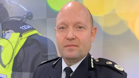 A shaven-headed man in formal police uniform standing against a backdrop depicting police work and graphics.