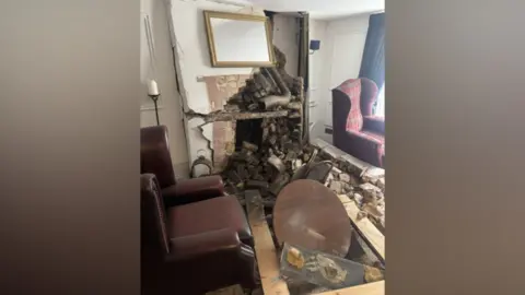 Averil Phillips & Family The offices of a funeral directors, with bricks all fallen out of a wall. Two chairs and a table are knocked over, with other damage around the room. There is a mirror on the wall which appears dislodged.