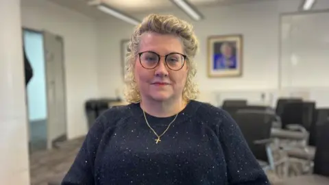 Lisa is sitting down, a blurry office setting is visible in the background. She is wearing a navy sparkly jumper, black rimmed glasses and has curly blonde hair. 