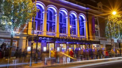 A building with the words Grand Theatre on it is lit up at night with people walking by outside. Two trees with fairy lights on are either side of it, along with a bright street lamp. It is nighttime.
