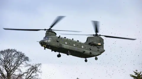 Chinook flying low to the ground just above the tree-line with debris swirling about.
