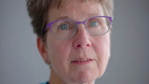 Judith Gibbons has short light brown hair, bright sea-blue eyes and purple rimmed glasses.