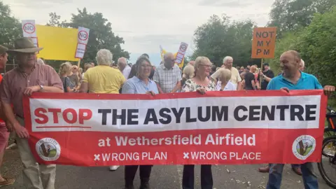 Los residentes de Mousumi Bakshi/BBC cerca de Wethersfield en una carretera sosteniendo una pancarta y carteles.