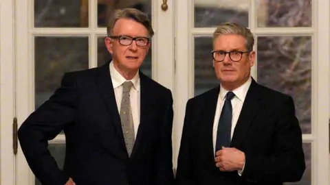 Carl Court/PA Lord Mandelson and Sir Keir Starmer are stood in front of two white framed, glass-panelled doors. They are staring straight ahead and both wearing dark coloured suits, white shirts and patterned ties.