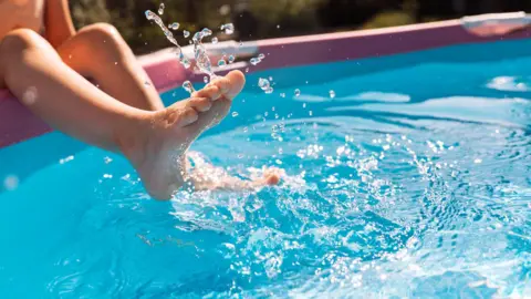 Getty Images A child splashing water with their feet in a pink inflatable pool on a sunny day. Only the legs are visible as droplets fly into the air, creating a joyful and carefree summer scene. The clear blue water contrasts playfully with the pink pool walls.