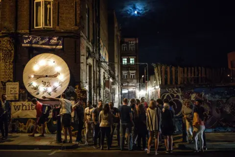 Getty Images A large crowd gathers on the street in the middle of the night with the graffitied buildings sitting in the background.