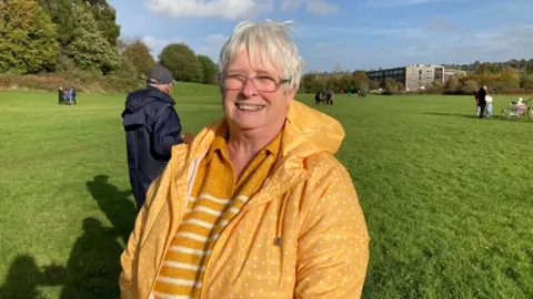 Martin Heath/BBC Liz Scott with short white hair and glasses wearing a yellow raincoat and standing on grass