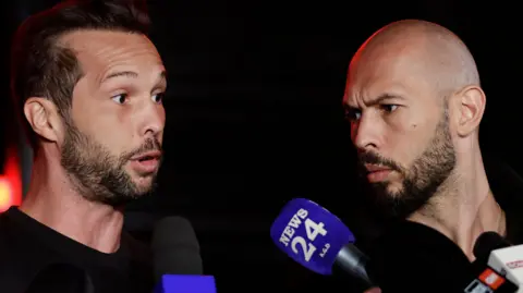 Robert Ghement/EPA-EFE/REX/Shutterstock Tristan Tate has a dark beard and dark hair with a receding hairline. He is speaking to the media and two microphones are in the shot. Andrew Tate has a dark beard and a shaved head and is to the right of the image, watching his brother.