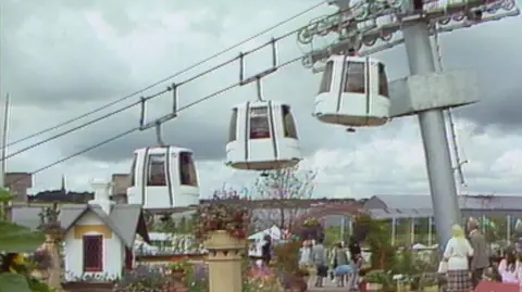 Three cable cars can be seen, side-on, climbing above a festival with people walking around and ornamental garden. The cable car gondolas are white, with a large pillar sticking up out of the ground to hold the cables.
