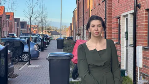 Eric Johnson/BBC A woman in a dark green top stands outside her home in a street. There are parked cars and bins behind her.