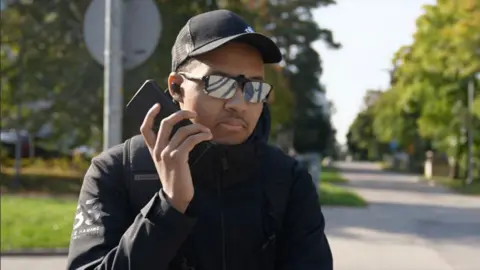 Photo of Charles-Emmanuel Mikko Rasanen, wearing a black jacket, parasols and a black cap, holding a mobile phone on a road near his home in Finland