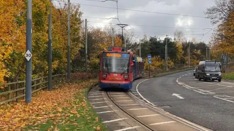 Oli Constable/BBC A red and blue tram traveling along tracks that run parallel to a road on the right side. The tram displays a digital sign reading “Middlewood.” A black car is driving on the road beside the tram tracks.
