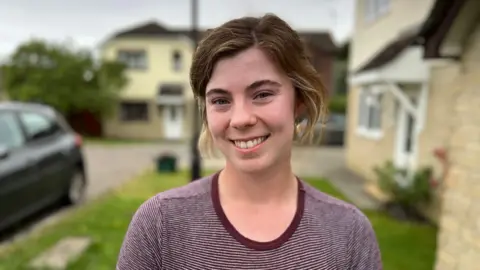 Women with purple and achromatic  lined apical  on, smiling astatine  the camera with blurred inheritance  of houses and greenish  grass.