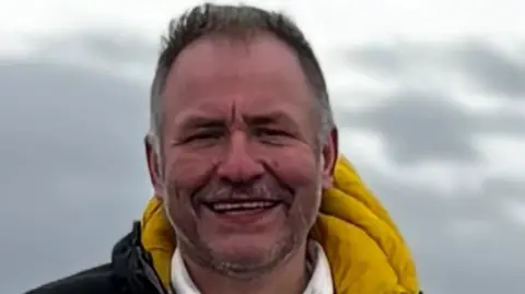 Alan Bolton stands outside where there are grey clouds in the background. Alan has short grey hair and stubble. He is wearing a black and yellow coat. 