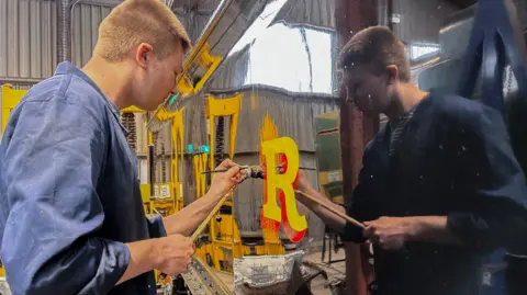 A man painting the letter 'R' onto the side of a locomotive in a works shed