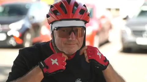Kevan Collins is putting a red bike helmet on to his head. He has matching red fingerless cycling gloves and is wearing a black top. He is also wearing large sunglasses on his face.